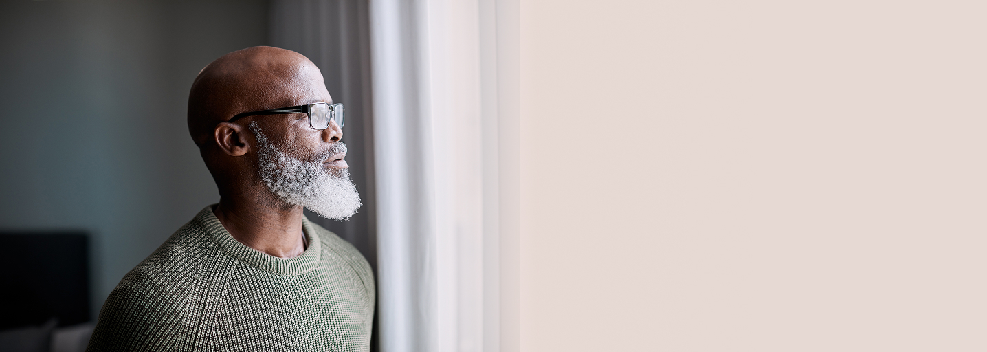 A person of color ,wearing glasses and a green sweater, stands by a window with white curtains. The person has a bald head and a white beard. The background is out of focus.